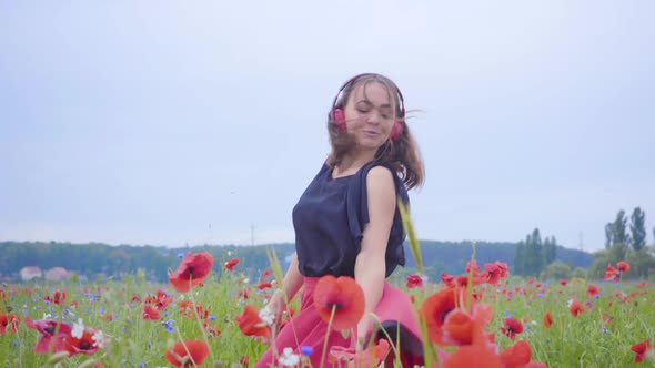 Cute Young Woman Wearing Headphones Listening To Music and Dancing in a Poppy Field Smiling Happily alt