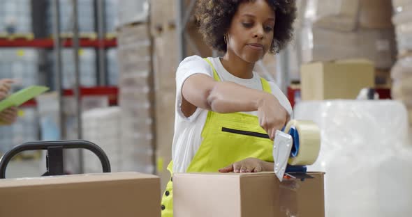 African Female Worker Packing Cardboard Box with Tape Gun Dispenser in Warehouse alt