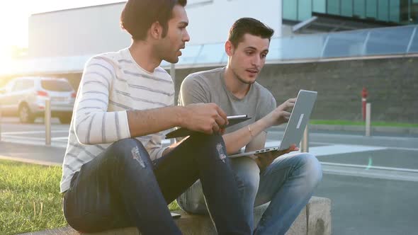 two young multiethnic men sitting outdoor using digital tablet and personal computer alt