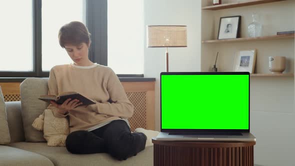 A Laptop with a Green Screen Stands on a Table Against of a Woman Reading a Book alt