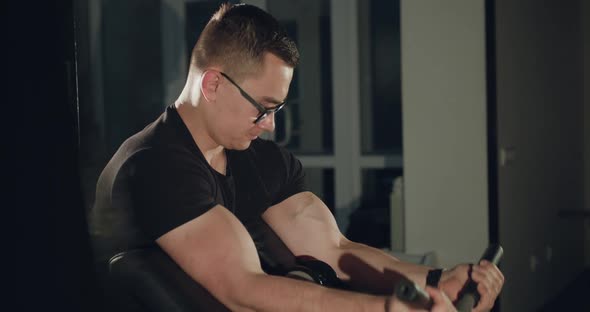 Athlete with Glasses Works Out with the Simulator to Pump Biceps in a Gym alt