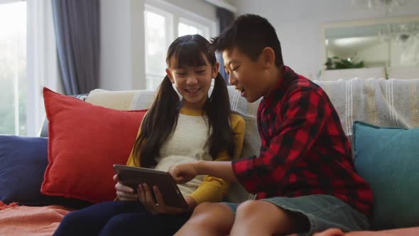 Asian brother and sister smiling and using tablet sitting on sofa at home alt