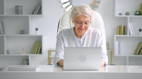 Smiling aged woman in glasses looks at laptop keyboard alt