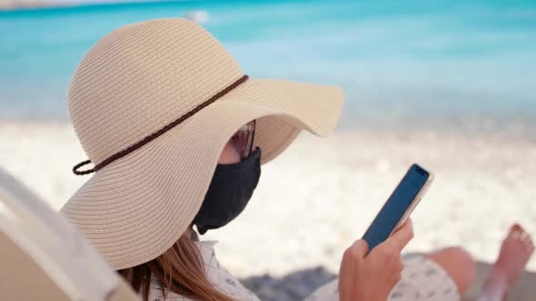Coronavirus Vacation: Woman in Face Mask Uses Phone App on Sea Beach in Summer
