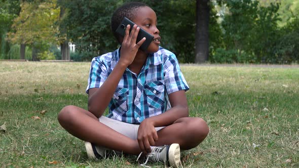 A Young  Black Boy Sits on Grass in a Park and Talks on a Smartphone alt