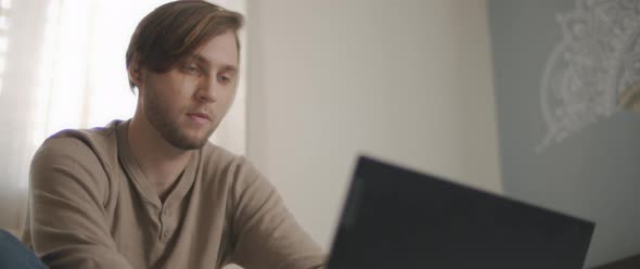 A close up of a young man sitting in bed and working on a computer.  alt