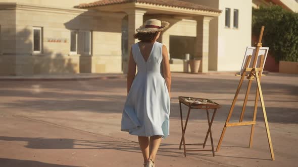 Live Camera Follows Confident Young Female Painter Walking in Slow Motion to Easel Taking Paint alt