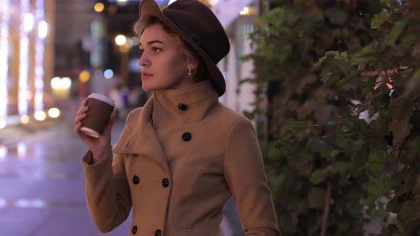 Beautiful Woman in Hat and Coat Drinks Coffee in the Night City