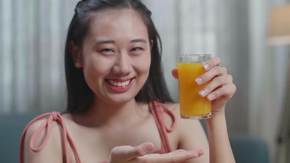 Happy Asian Woman Holding And Pointing A Glass Of Orange Juice While Having Healthy Food At Home alt