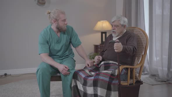 Middle Shot of Old Caucasian Man Sharing with Adult Male Nurse. Young Guy Taking Hand of Wise Senior alt