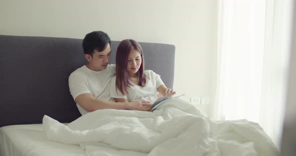 Asian husband and pregnant wife spend time together in the bedroom and reading a book to a child.