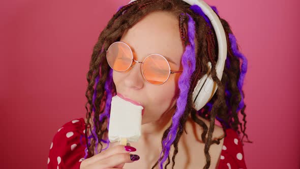 Young beautiful woman in bright sunglasses, wireless white headphones licking ice cream alt