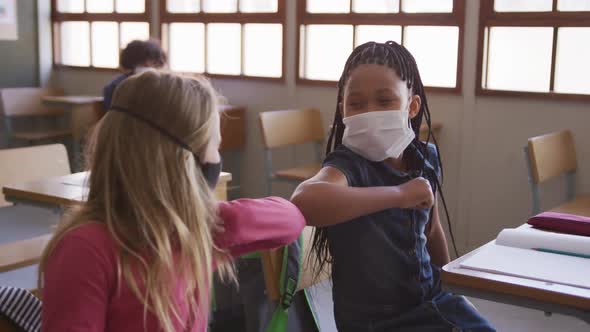 Two girls wearing face masks greeting each other by touching elbows at school alt