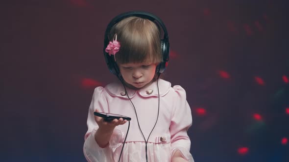 Little Girl in Big Headphones and Vintage Dress is Listening to Music on Phone alt