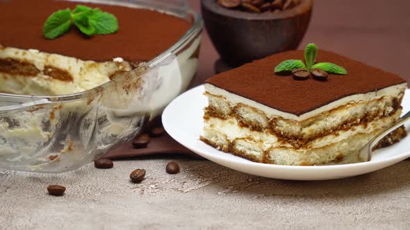 Portion of Traditional Italian Tiramisu dessert and baking dish on grey concrete background alt