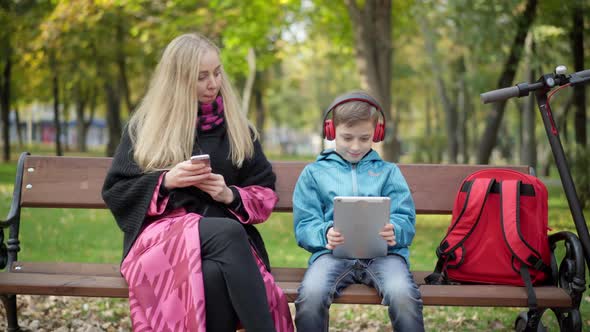 Absorbed Caucasian Young Mother and Little Son Surfing Internet on Smartphone and Tablet Sitting in alt