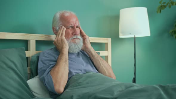 Tired Elderly Man Has Acute Headache While Sitting on Ahe Bed At Home alt