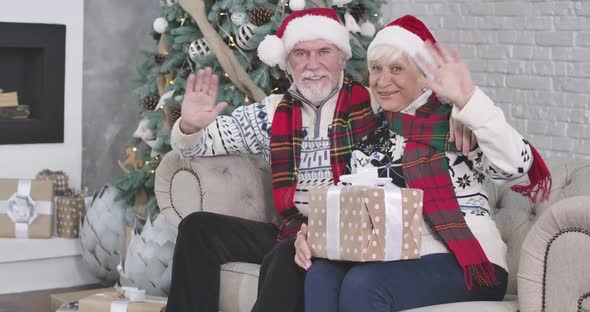 Medium Shot, Smiling Old Caucasian Man and Woman Sitting on Couch with Presents and Waving at Camera alt