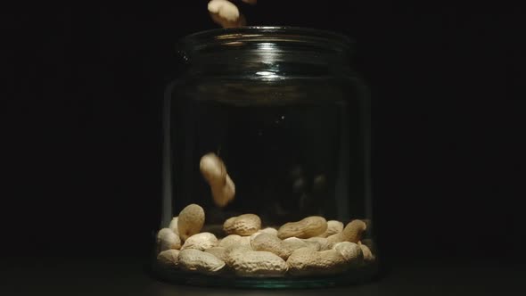 SLOW MOTION: Unpeeled Peanuts Fall Into Empty Glass Jar On Black Background - Front View alt