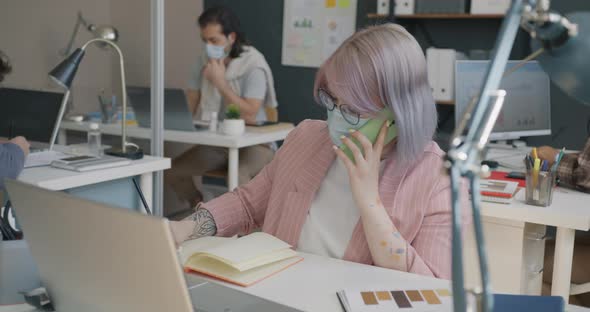 Female Employee Wearing Face Mask Chatting on Mobile Phone Indoors at Workplace alt