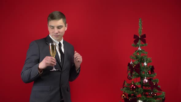 Young Man Dancing with a Glass of Champagne Near the Christmas Tree on a Red Background. A Man in a alt