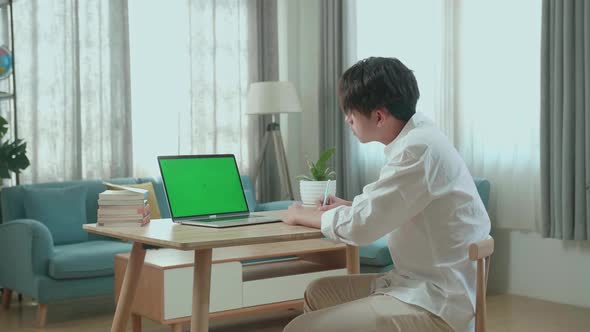 Asian Man Student Look At Green Screen Computer And Write In The Notebook While Studying Online alt