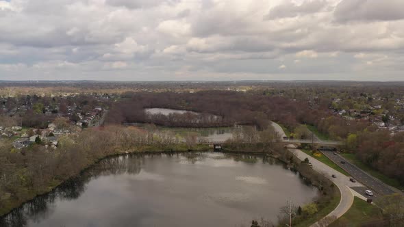 A high angle drone view over a quiet lake, surrounded by dry, brown trees and a road. It is a cloudy alt