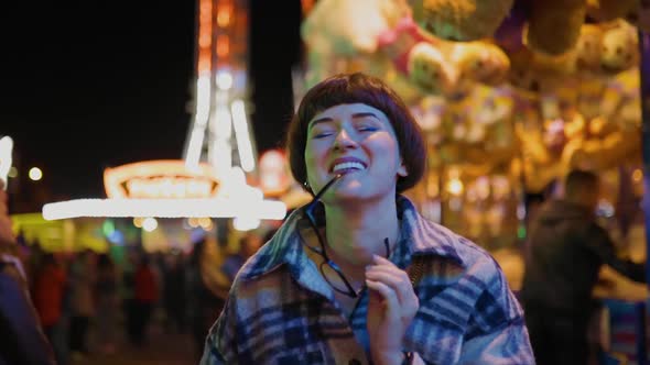 Young Woman Holds Glasses in Teeth at Amusement Park with Bright Lights alt