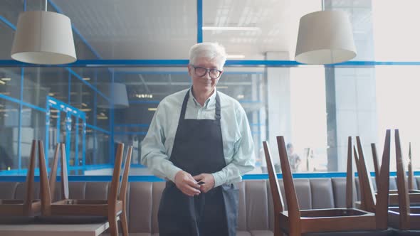 Mature Small Business Owner Arranging Apron Walking in Closed Cafe alt