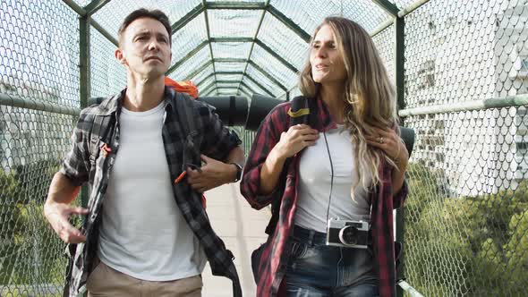 Couple of Travelers with Backpacks Walking on Bridge From City