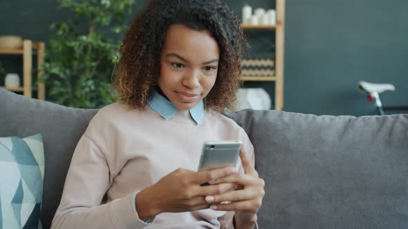 Attractive Afro-American Girl Holding Smartphone Touching Screen Relaxing at Home alt