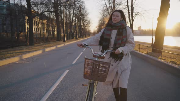 Brunette Woman in Scarf Carries Yellow Bicycle on City Road alt
