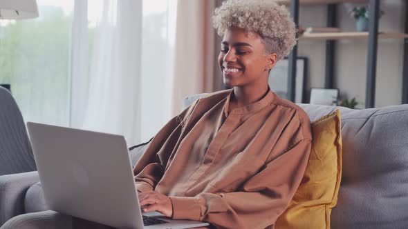 Happy Young African Woman Sitting on Couch Using Laptop Looking at Screen alt