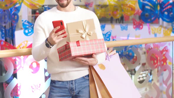 Attractive Man in Shopping Mall with Christmas Gifts in Hand Uses Smartphone alt