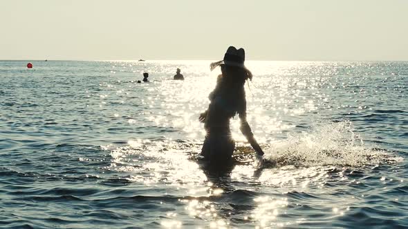 Silhouette Of Girl In Fashionable Hat Playing On The Sea Water. Sun Flare alt