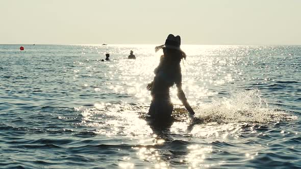 Silhouette Of Girl In Fashionable Hat Playing On The Sea Water. Sun Flare