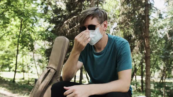 Young Man Putting on Face a Medical Mask While Is Sitting in the Park alt