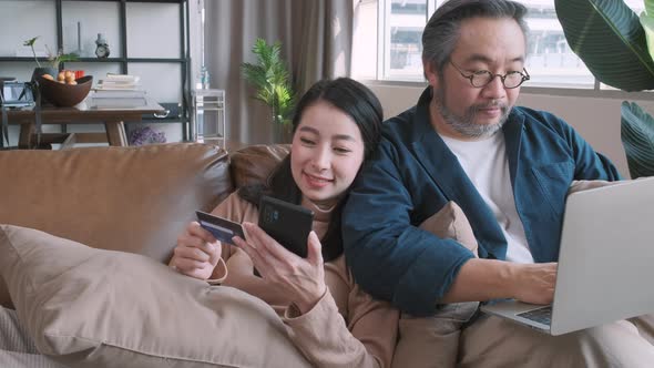 Man looking at his wife shopping online by smartphone and credit card in living room at home