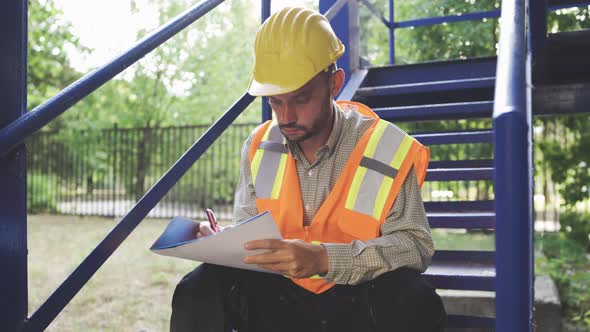 Civil Engineer in Helmet and Safety Vest Taking Notes in Notepad alt