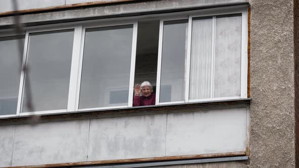 Elderly Woman Waves Hand Goodbye Greeting From Window Of Balcony On Isolation alt