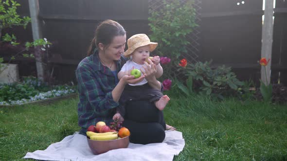 Happy Young Cheerful Mother Holding Baby Eating Fruits On Green Grass