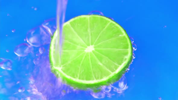 Water pours on green hue juicy lime on blue background slow motion closeup. Lime slice water alt