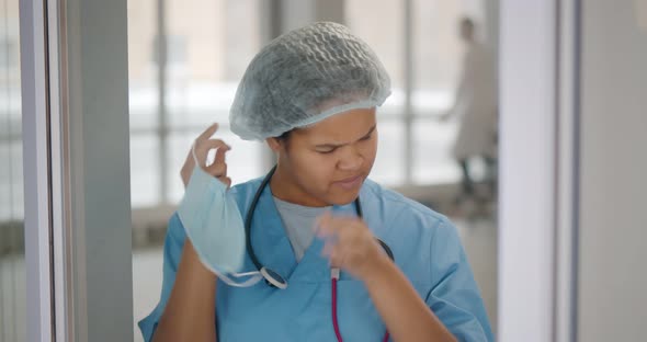 Tired and Exhausted Female African Scrub Nurse Removing Face Mask and Standing Near Window alt