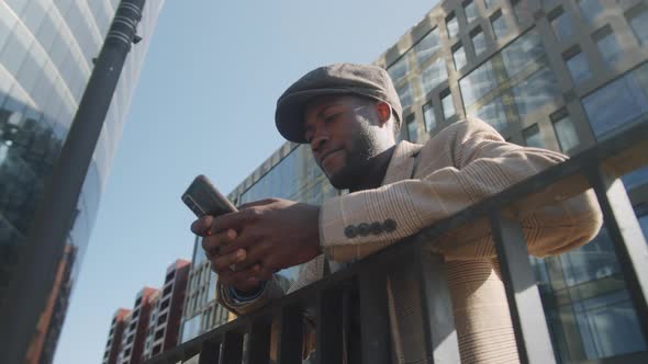 Black Businessman Leaning on Railing and Typing on Smartphone alt