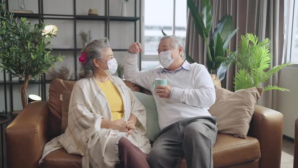 asian retired couple wear face mask sit relax on sofa couch conversation together