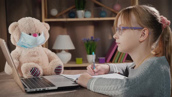 School Online  Pupil Doing Lessons Near Laptop Next to a Teddy Bear in a Protective Mask alt