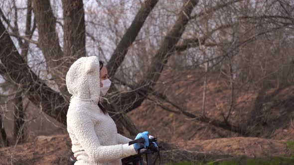 Young Woman in Medical Mask and Gloves Stands with Bicycle Holding on to Rudder in Countryside