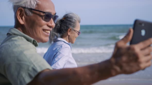 senior couple relaxing outdoors walking and taking selfie together with smartphone on beach alt