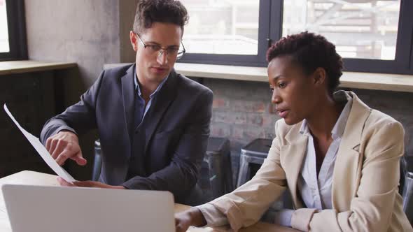 Diverse male and female business colleagues sitting talking in cafe looking at laptop and paperwork alt