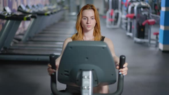 Front View Portrait of Concentrated Fit Caucasian Redhead Woman Riding Exercise Bike in Gym alt